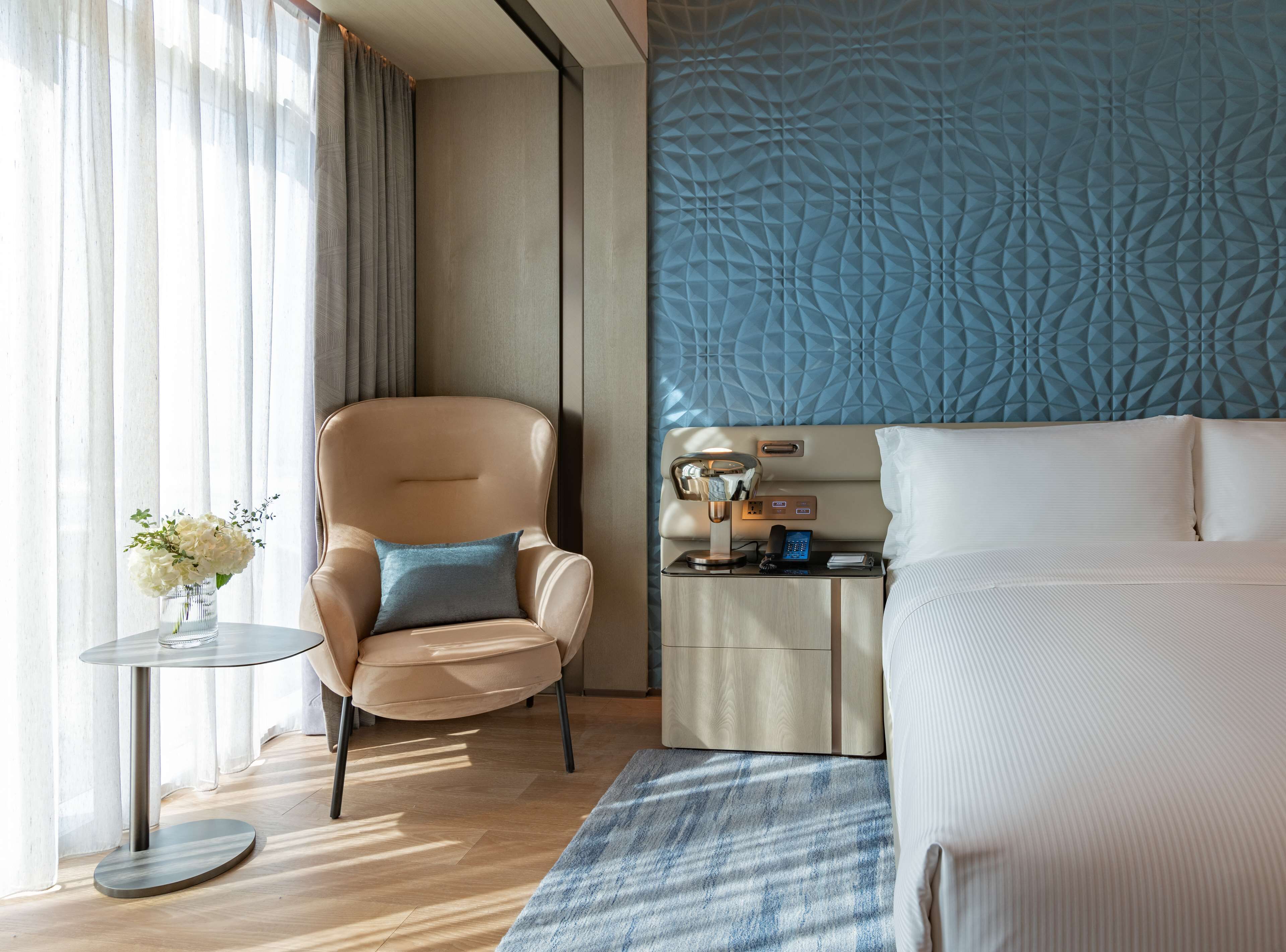 A hotel room with a bed, a chair, a small table with a vase of flowers, a nightstand with a lamp, and a wall with a geometric pattern.