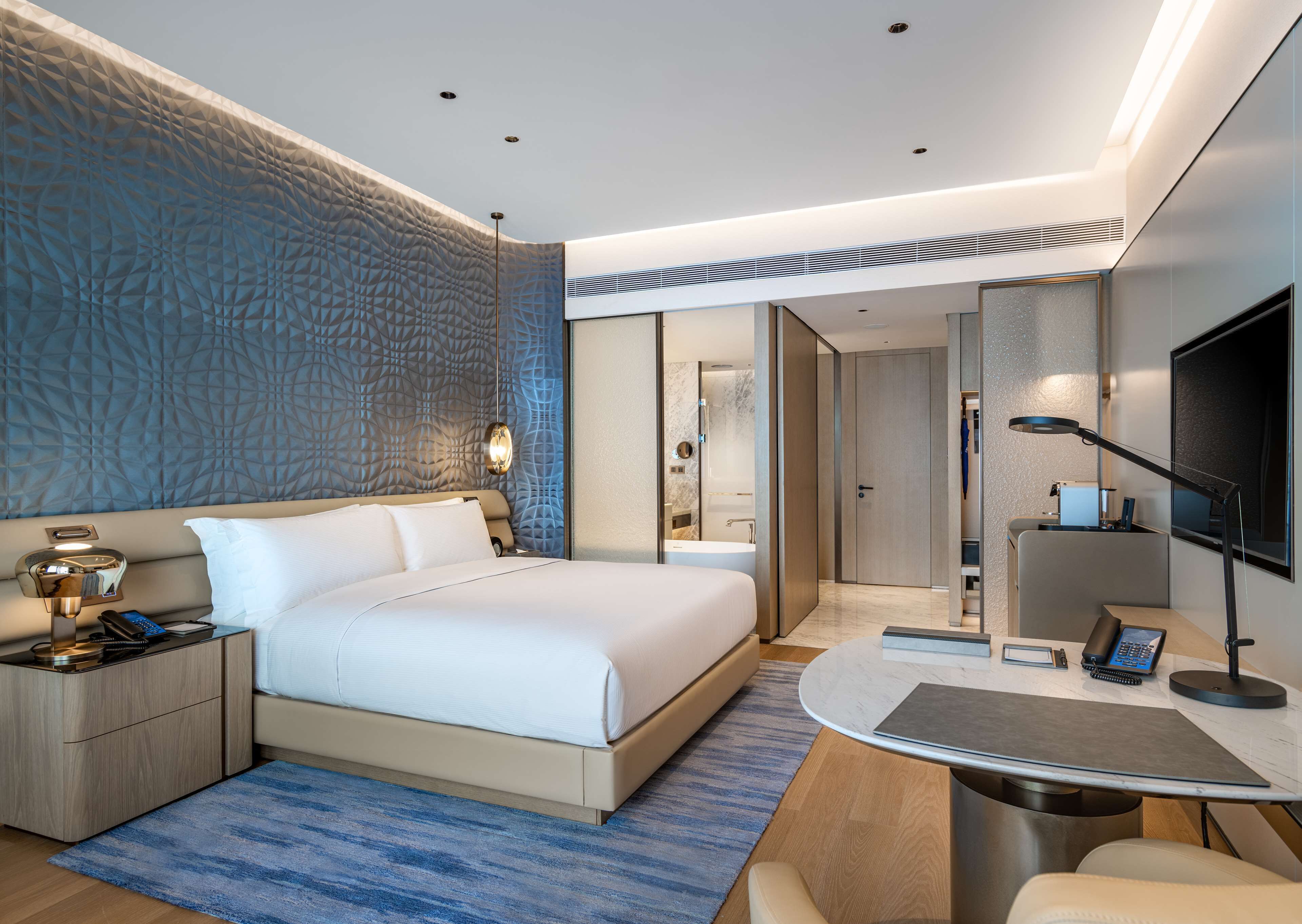 A modern hotel room with a large bed, a desk with a computer, a TV, and a bathroom visible through an open door.
