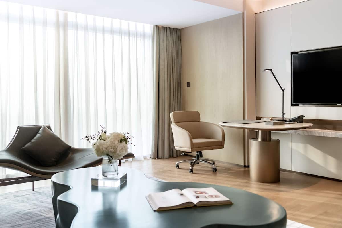 A modern hotel room with a curved sofa, a round table with a book, a chair, a TV, and a large window with curtains.
