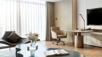 A modern hotel room with a curved sofa, a round table with a book, a chair, a TV, and a large window with curtains.