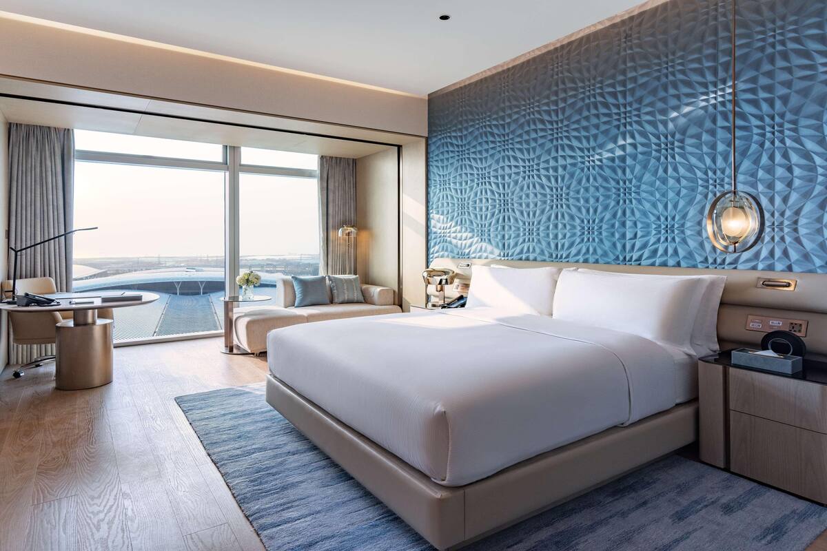 A modern hotel room with a large bed, a desk, and a view of a cityscape through large windows.