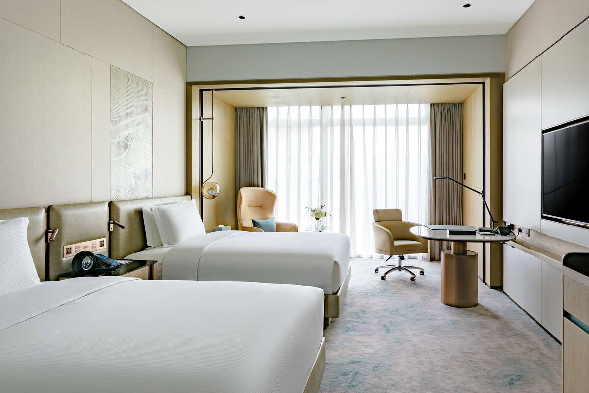 A hotel room with two beds, a desk, a chair, a TV, and a large window with curtains.