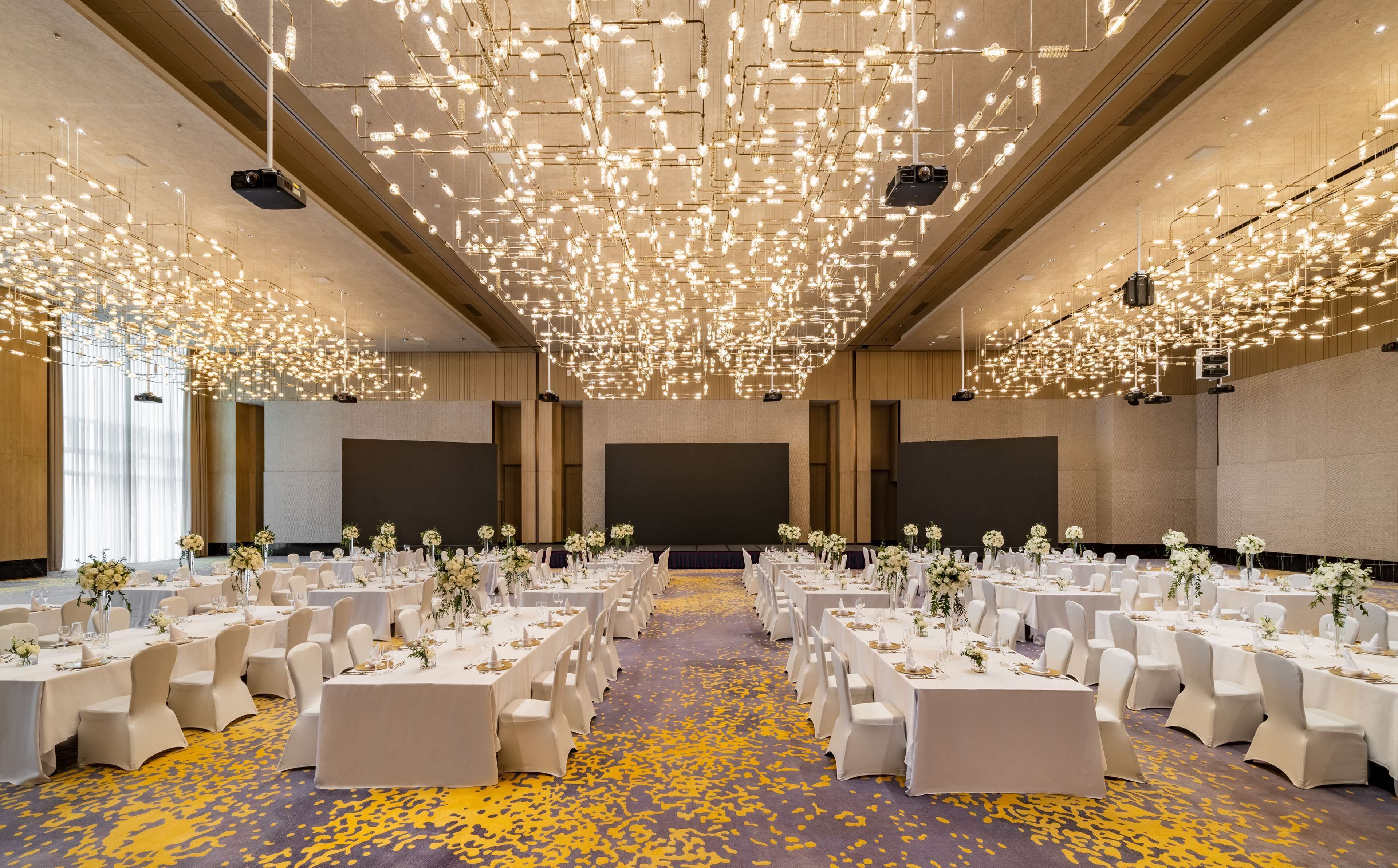 A spacious banquet hall with rows of tables set for events, featuring a modern chandelier and a carpeted floor.