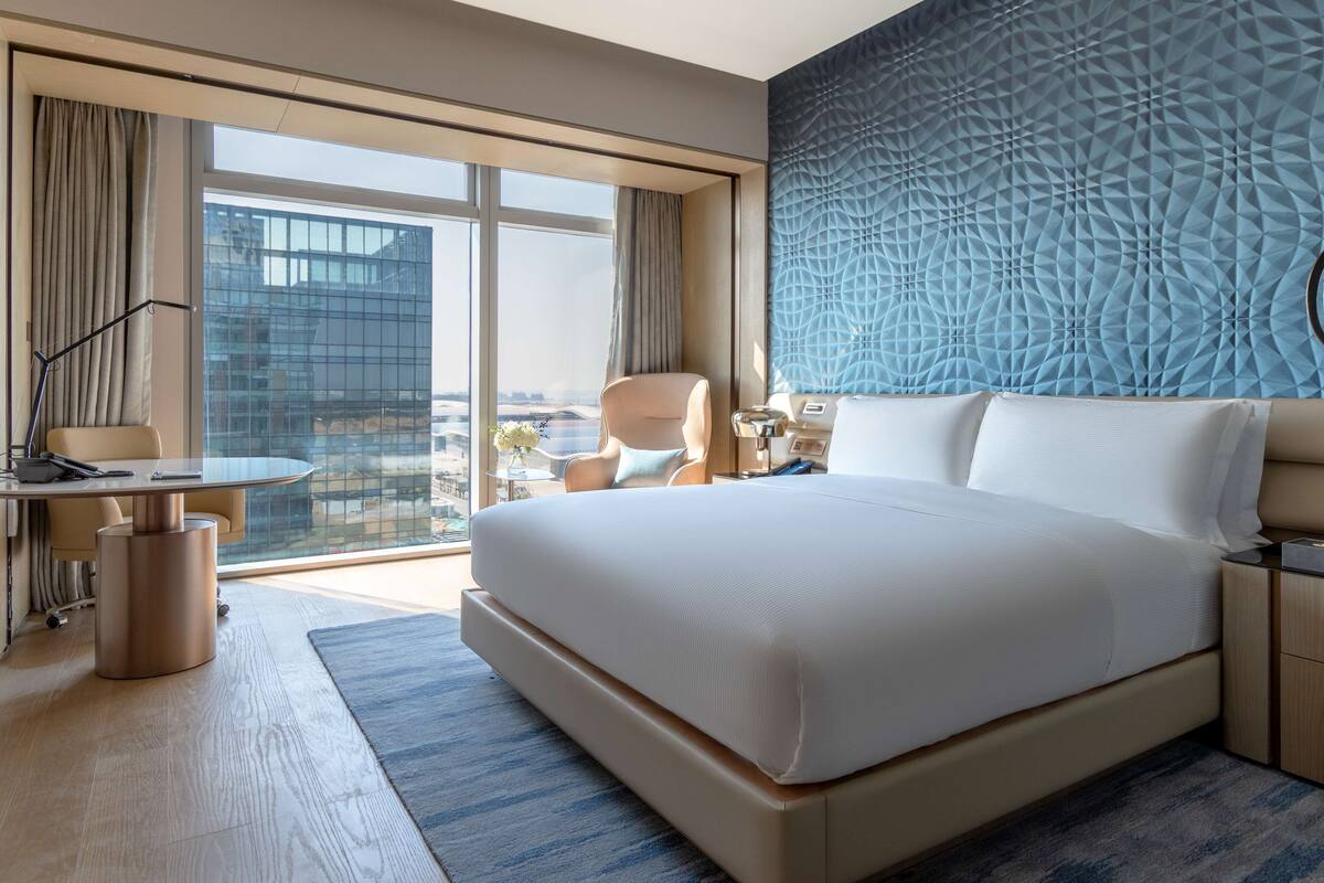 A modern hotel room with a large bed, a desk, and a view of a cityscape through floor-to-ceiling windows.
