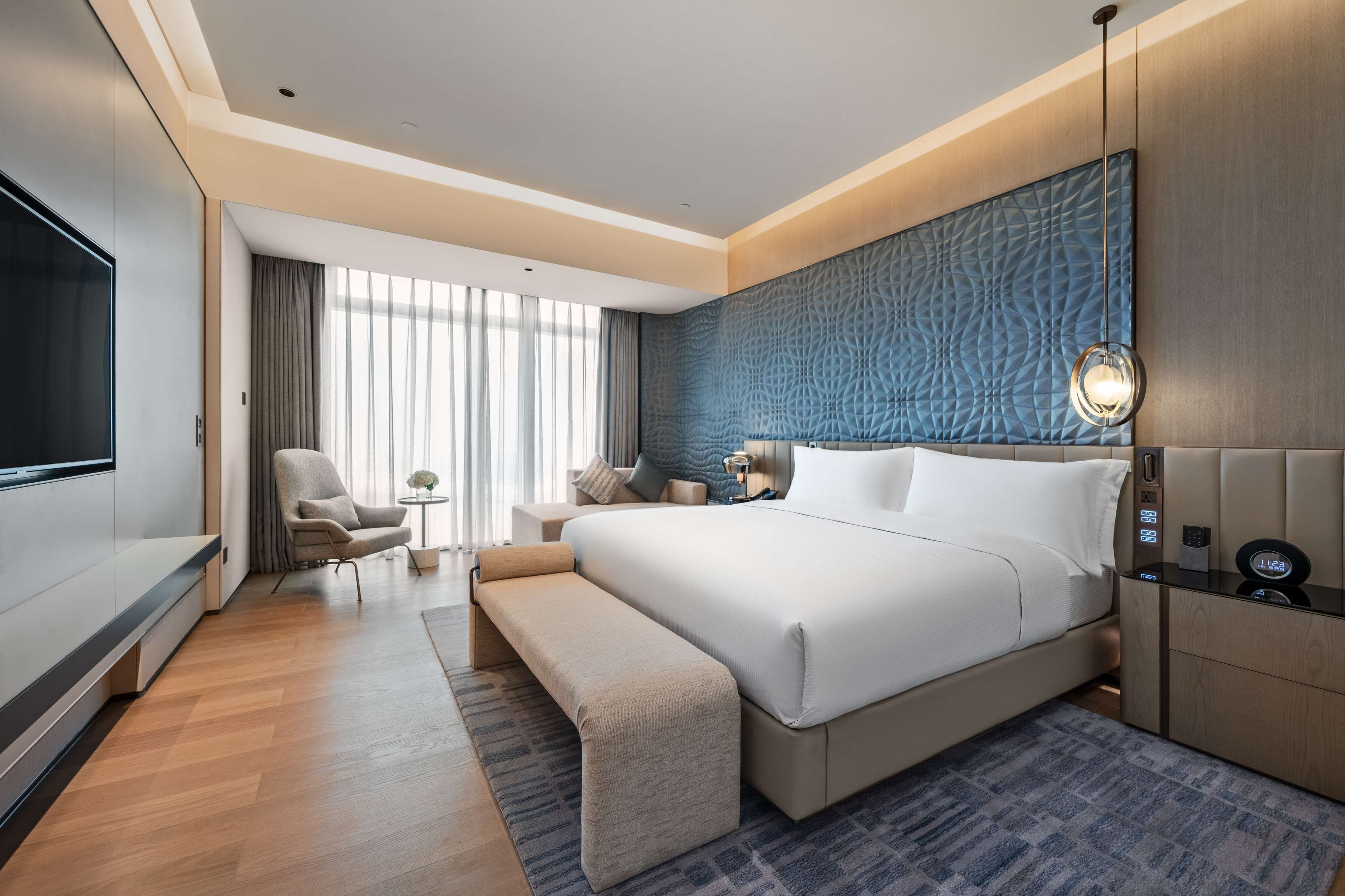 A modern hotel room with a large bed, a TV, a seating area with a chair and a small table, and a large window with curtains.