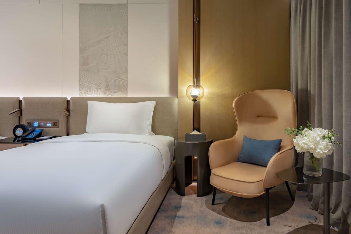 A hotel room with a bed, a chair, a lamp, and a small table.