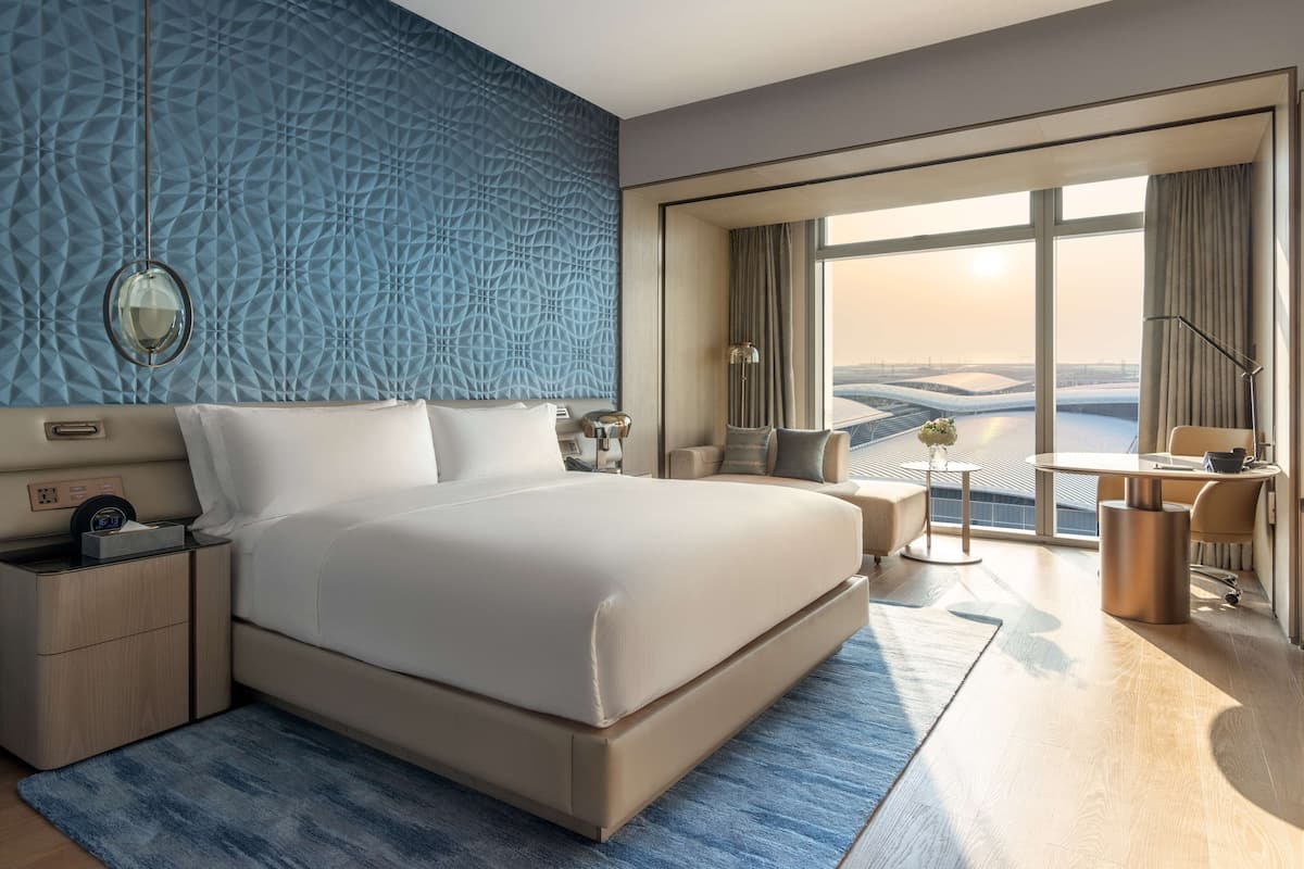 A modern hotel room with a large bed, a desk, and a view of a cityscape.