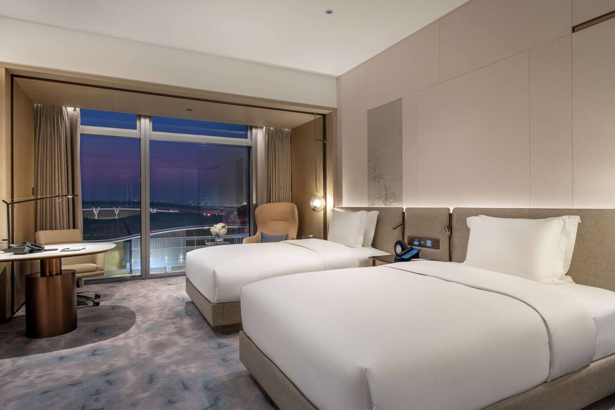 A hotel room with two beds, a desk, a chair, and a large window offering a city view.