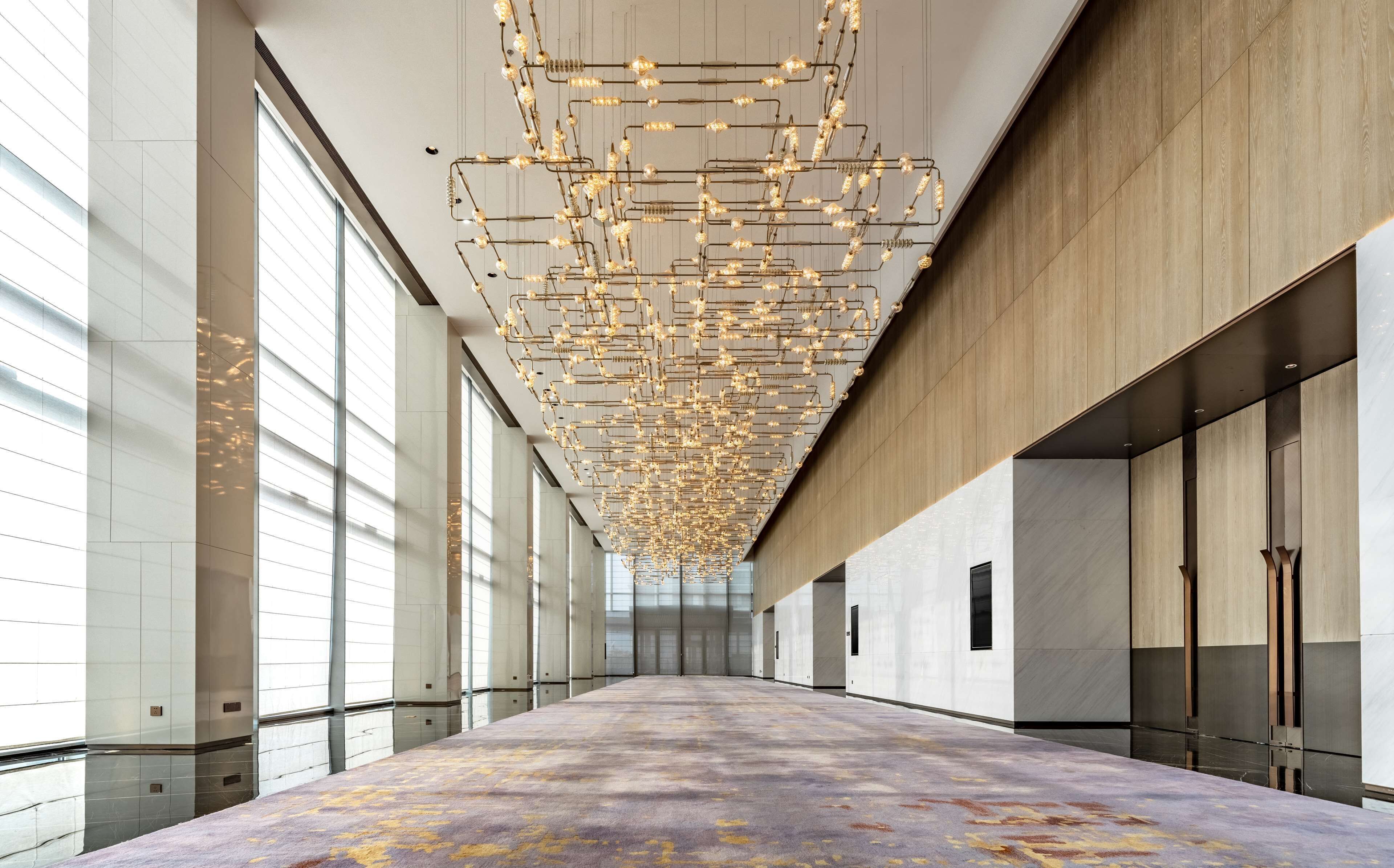 A spacious corridor with a patterned carpet, large windows, and a unique ceiling light fixture.