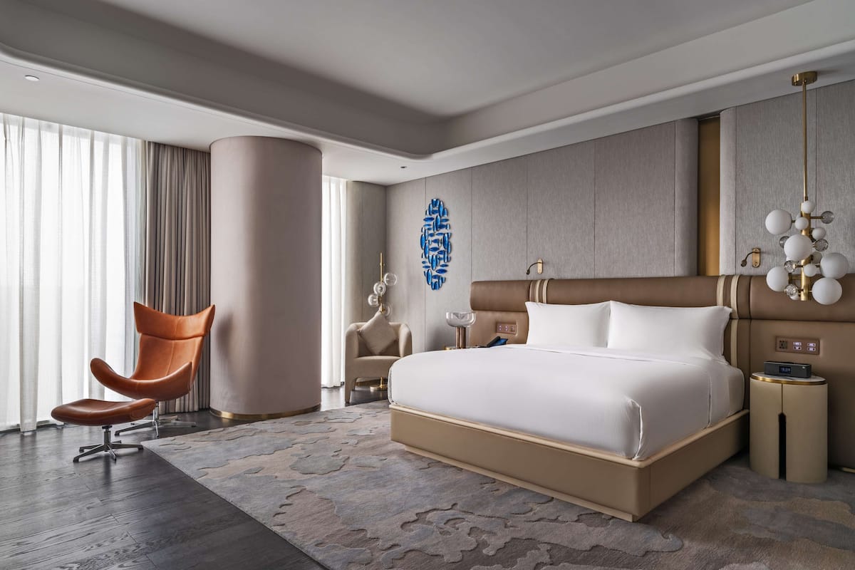 A modern hotel room with a large bed, a chair, a nightstand, and a decorative wall piece.