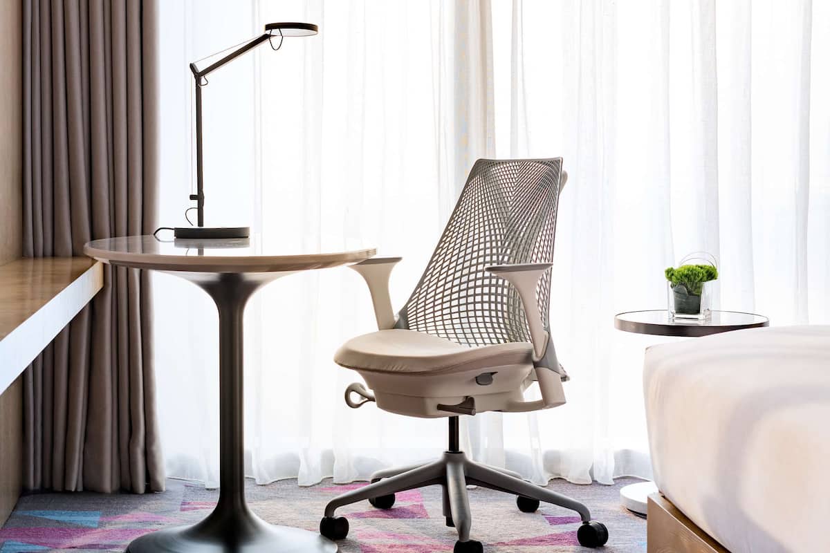 Een hotelkamer met een bed, een bureau met een lamp, een mesh bureaustoel en een kleine plant op een bijzettafeltje.