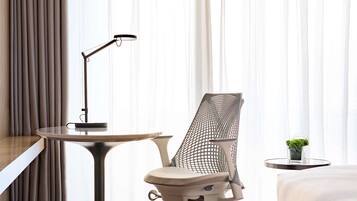 A hotel room with a bed, a desk with a lamp, a mesh office chair, and a small plant on a side table.