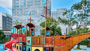 Een speeltuin met piratenthema, inclusief een glijbaan en klimconstructies, met moderne gebouwen op de achtergrond.