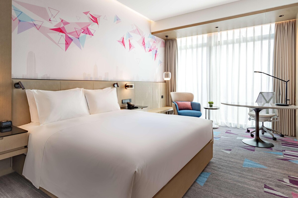 A hotel room with a large bed, a desk, a chair, and a cityscape mural on the wall.
