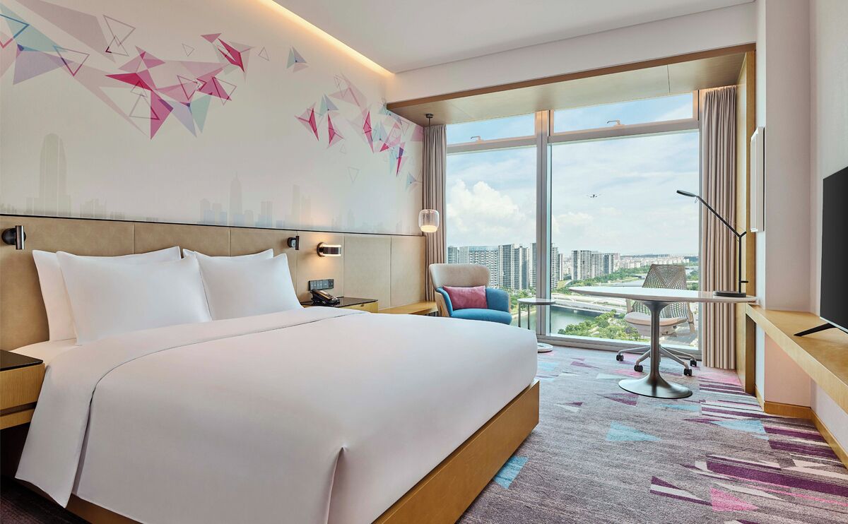 A hotel room with a large bed, a desk, a chair, a TV, and a view of the city through large windows.