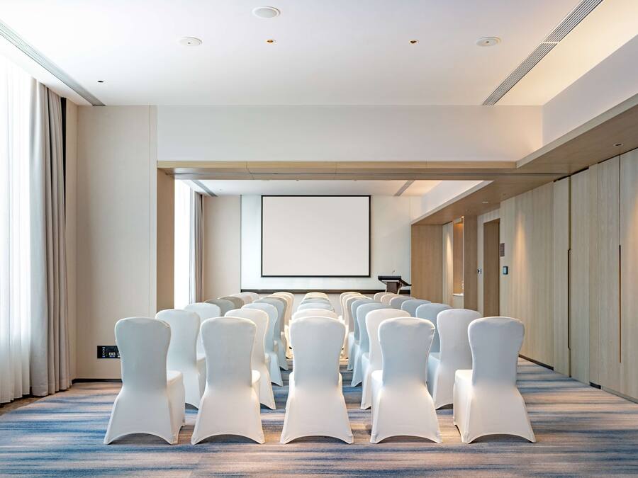 Un salón de conferencias con filas de sillas blancas, una pantalla grande y un atril.