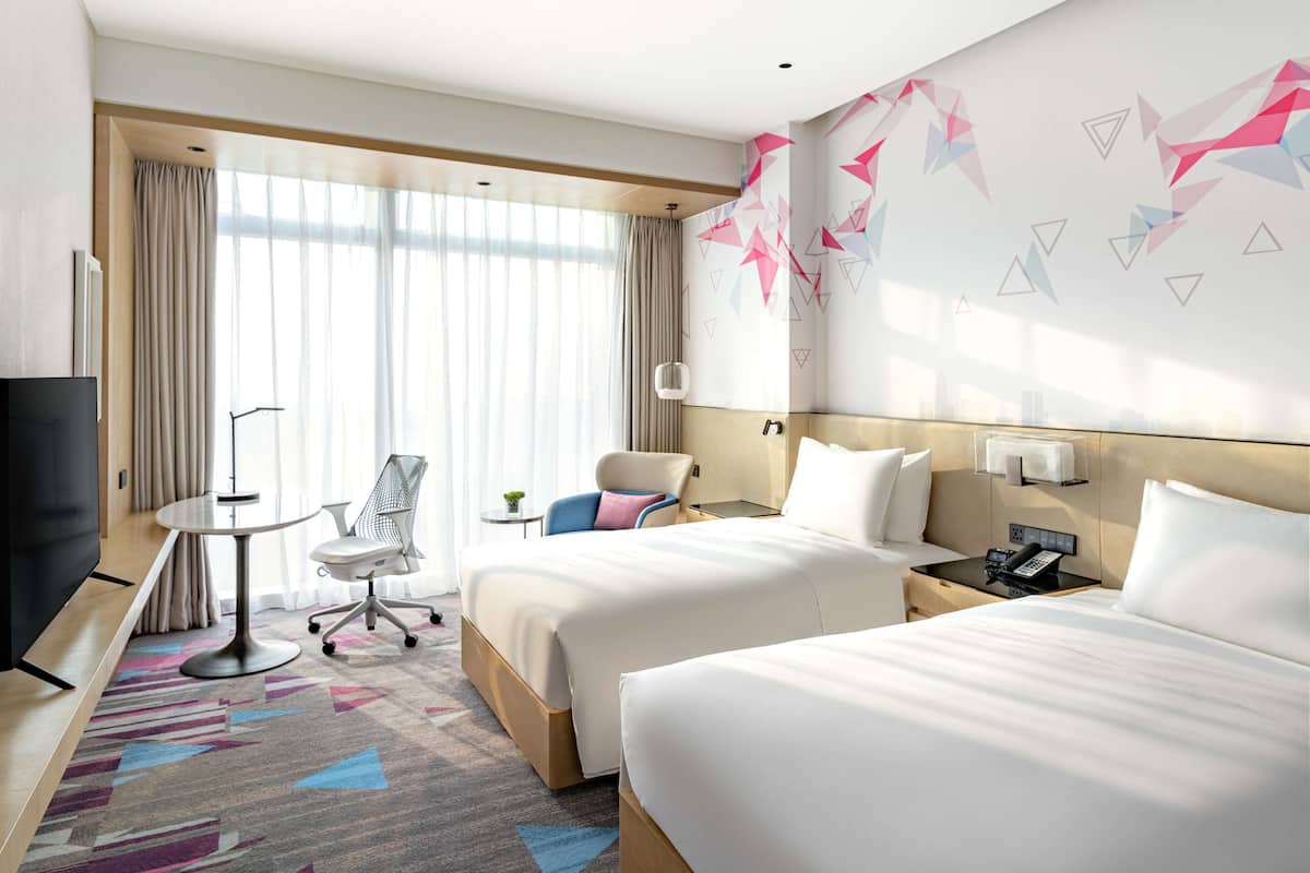 Een hotelkamer met twee bedden, een bureau met stoel, een televisie en een groot raam met gordijnen.