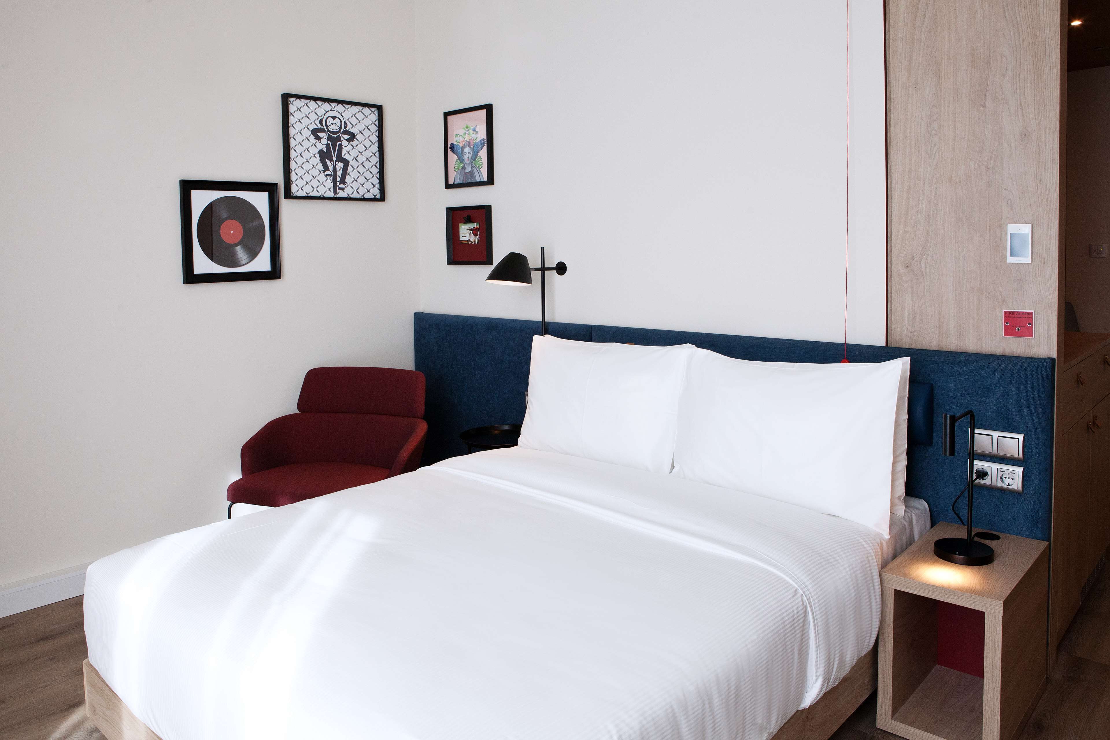 A hotel room with a bed, a red chair, a nightstand with a lamp, and framed artwork on the wall.
