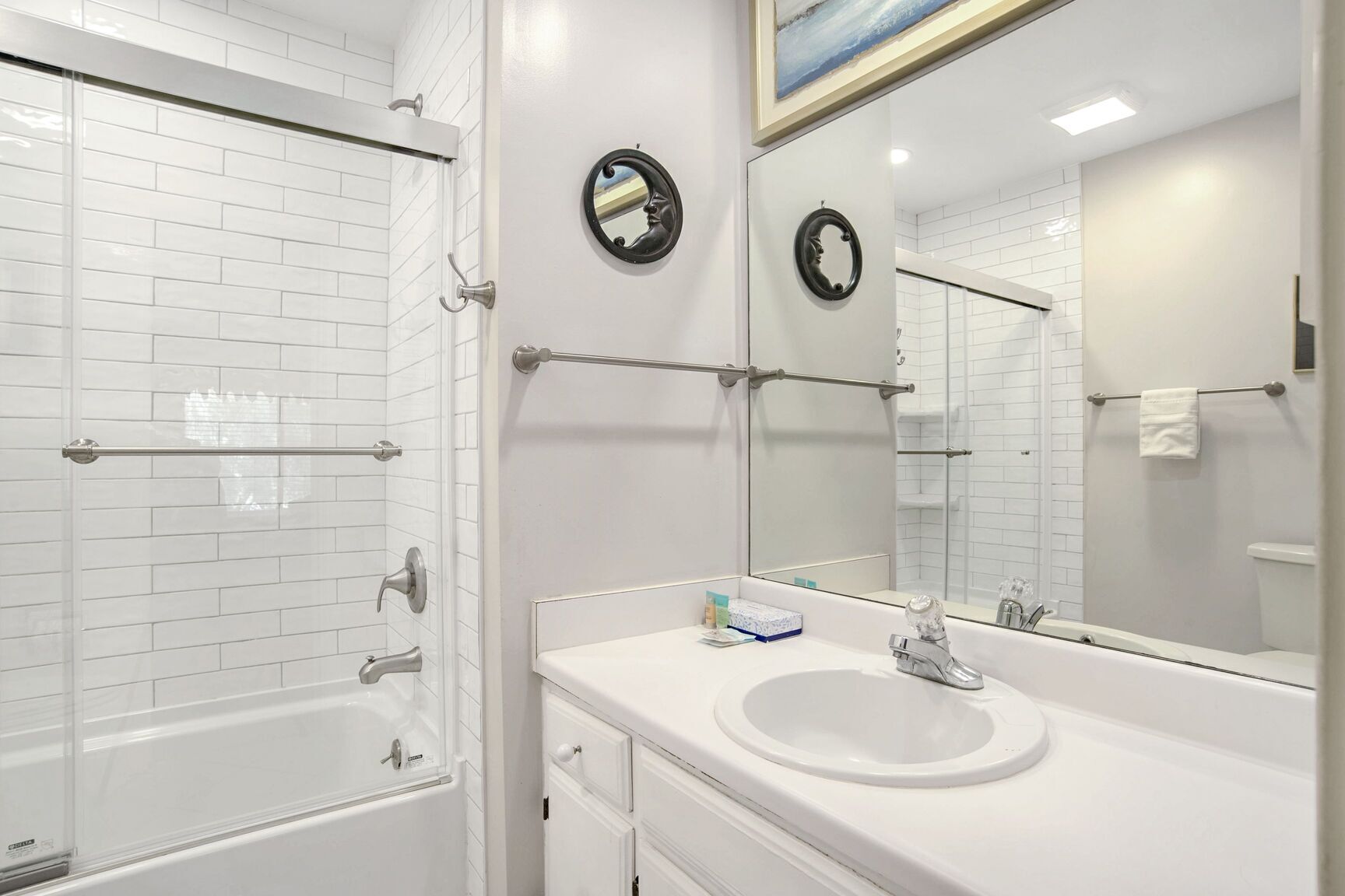 A modern bathroom with white tiled walls, a glass shower enclosure, a white sink with a mirror, and a towel rack.
