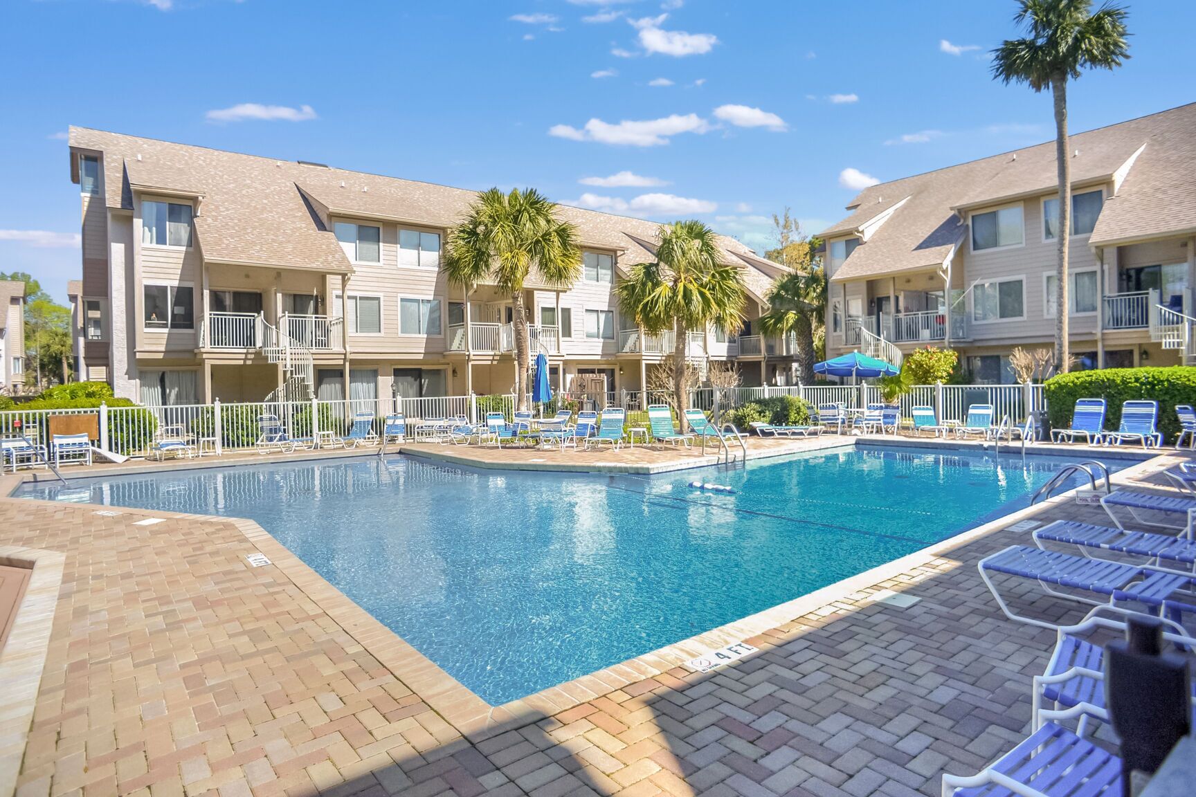 A swimming pool surrounded by lounge chairs and palm trees, with a brick walkway leading to the pool.