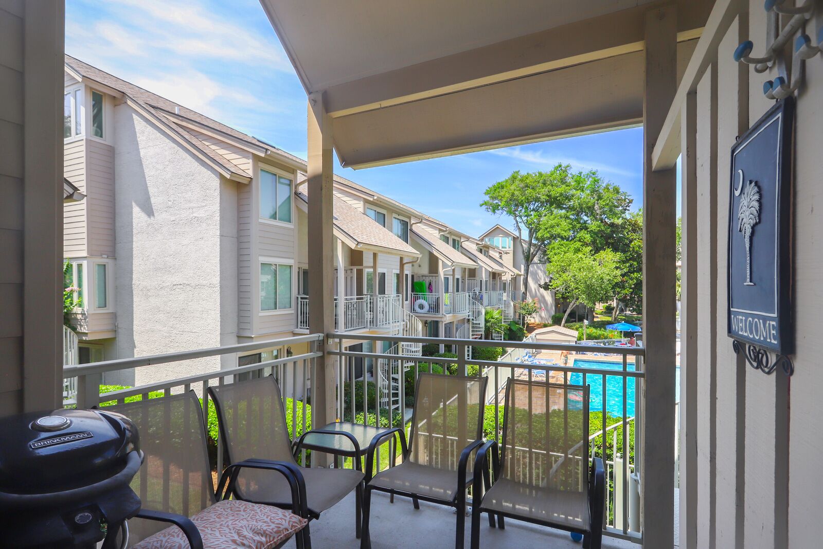 A balcony with a grill, chairs, and a view of a swimming pool and neighboring buildings.