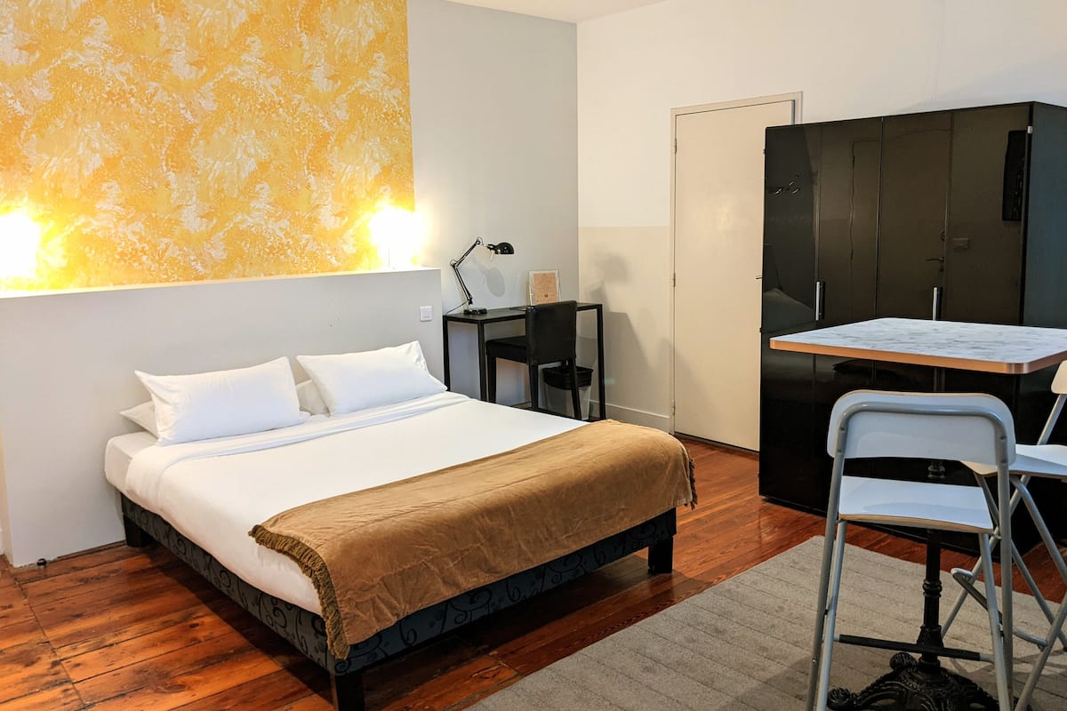 A hotel room with a bed, a desk with a chair, a wardrobe, and a wall-mounted lamp.