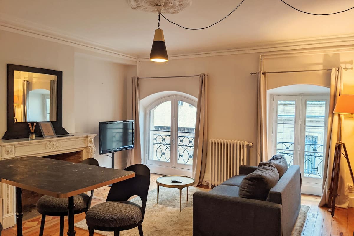 A living room with a sofa, a dining table with chairs, a TV, a small round table, and a balcony with a view.
