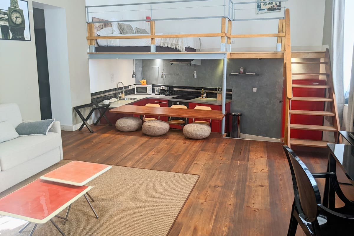 A compact studio apartment with a kitchenette, a dining area with a wooden table and stools, a bunk bed, and a staircase.