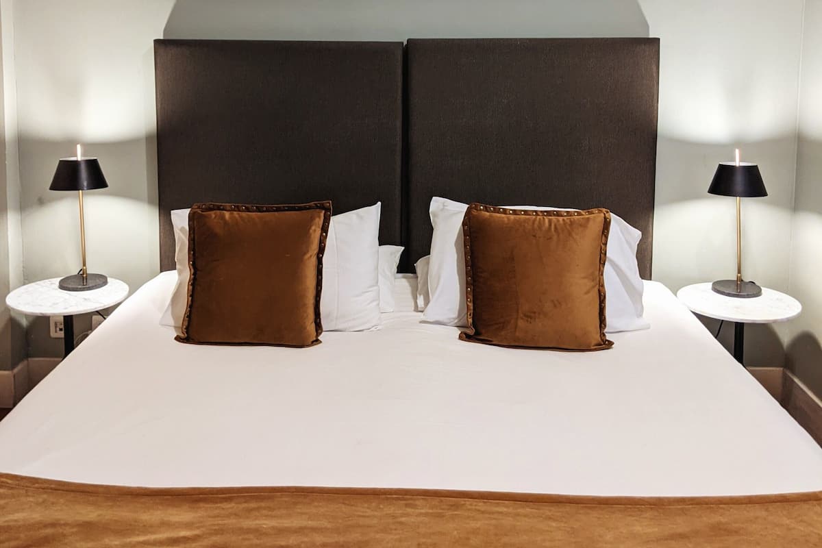 A neatly made bed with two brown pillows, a white comforter, and a large abstract painting above. There are two bedside tables with lamps on either side of the bed.