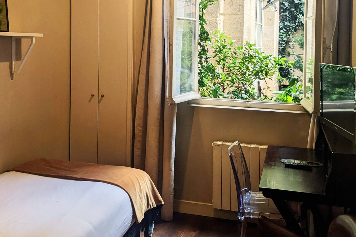 A bedroom with a bed, a desk, a radiator, and a window with a view of greenery.