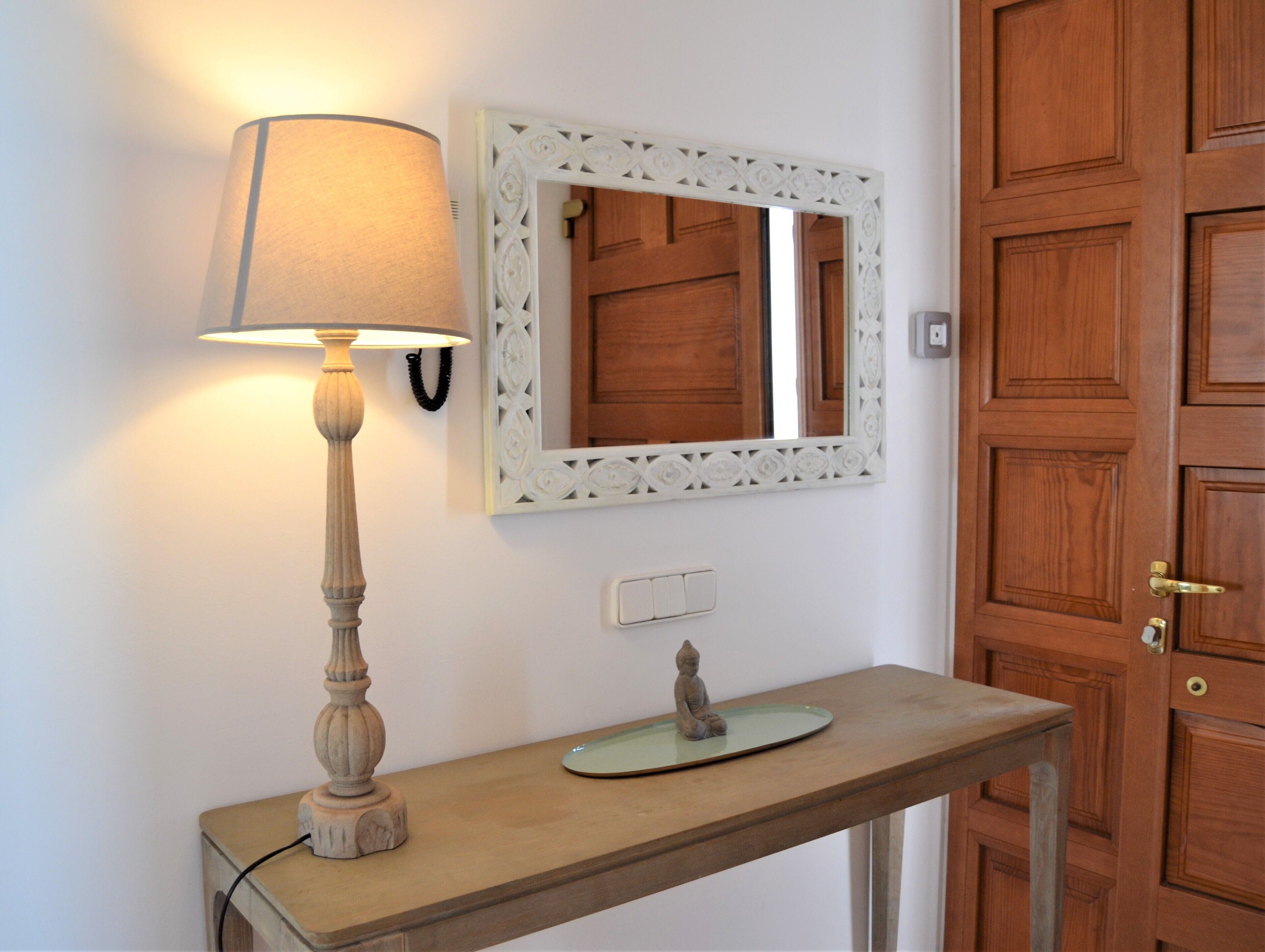 Una puerta de madera, un espejo con marco decorativo fijado a la pared, una mesa con lámpara y una pequeña estatua.