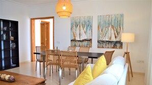 Een woonkamer met een bank, salontafel, eettafel en stoelen. Er hangen schilderijen aan de muur en staat een lamp.