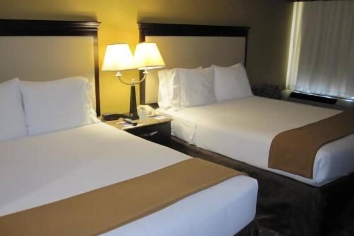 A hotel room with two beds, a nightstand with a lamp, and a window with curtains.