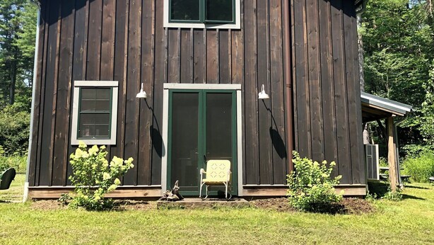 Kaksikerroksinen puutalo, jossa on etuovi, kaksi ikkunaa ja pieni kuisti, jossa on tuoli.