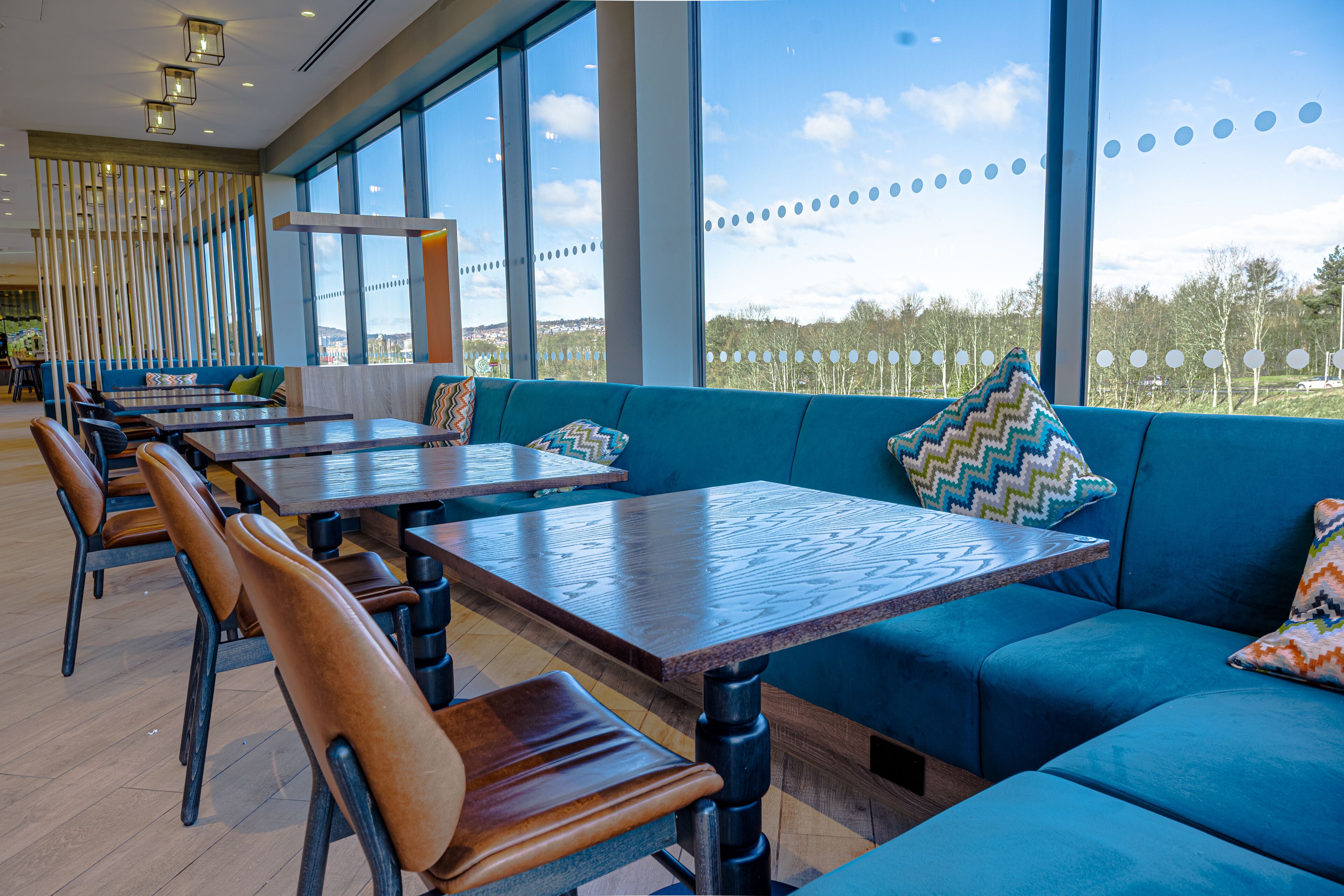 A restaurant with large windows, wooden tables, and blue upholstered seating.