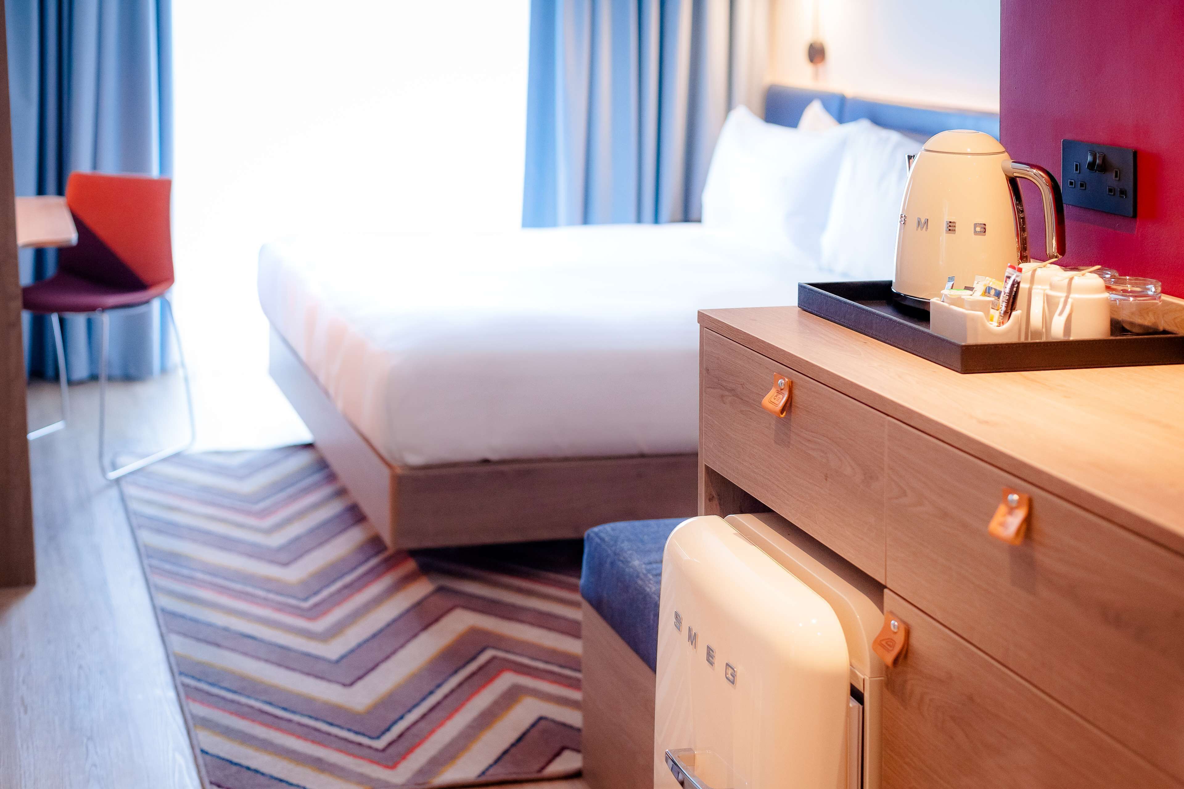 A hotel room with a bed, a wooden dresser, a coffee maker, and a small refrigerator.