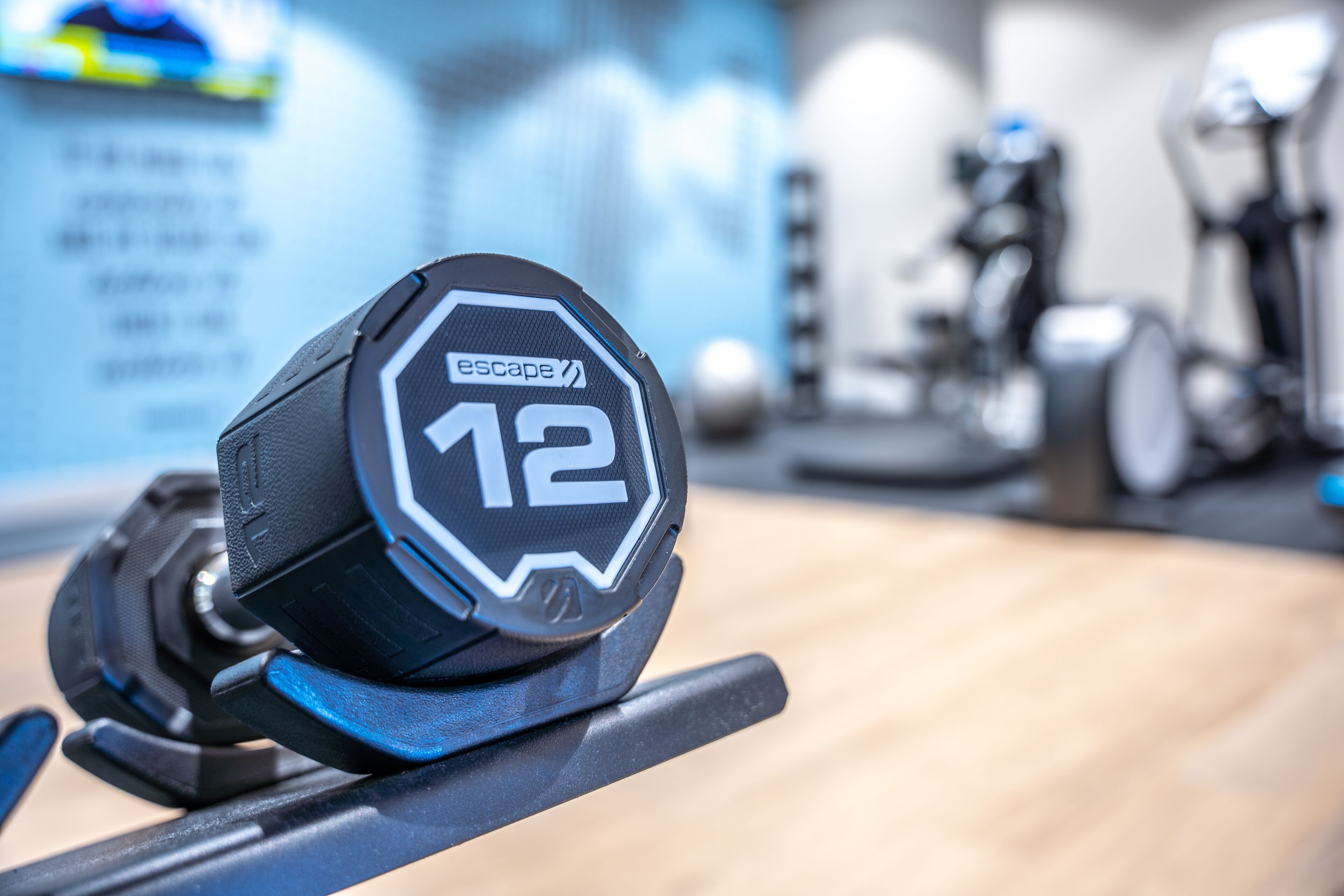 A weightlifting dumbbell with the number 12 and the word "escape" on it, placed on a rack in a gym.