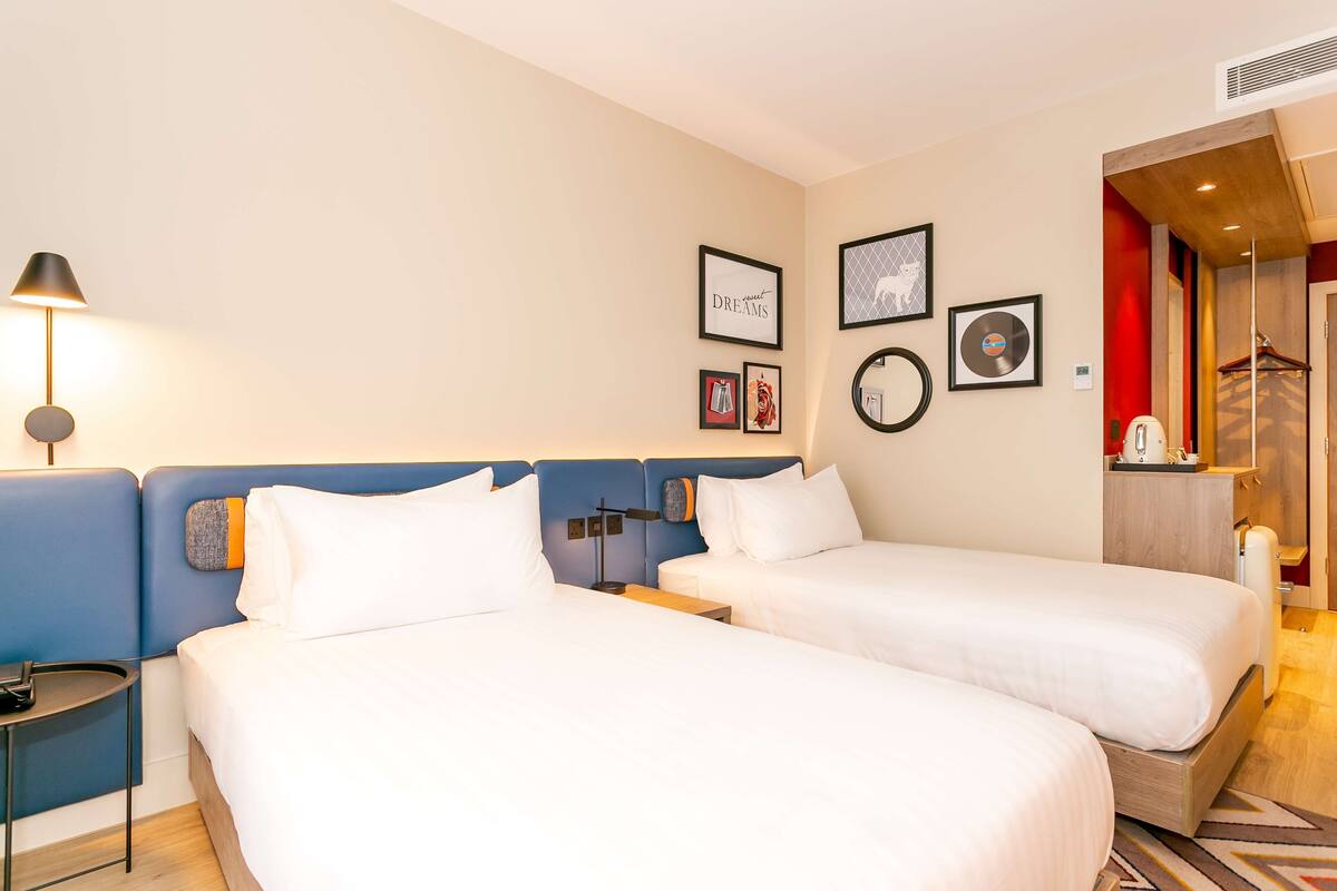 A hotel room with two beds, a bedside table, a lamp, and wall decorations including framed pictures and a CD.