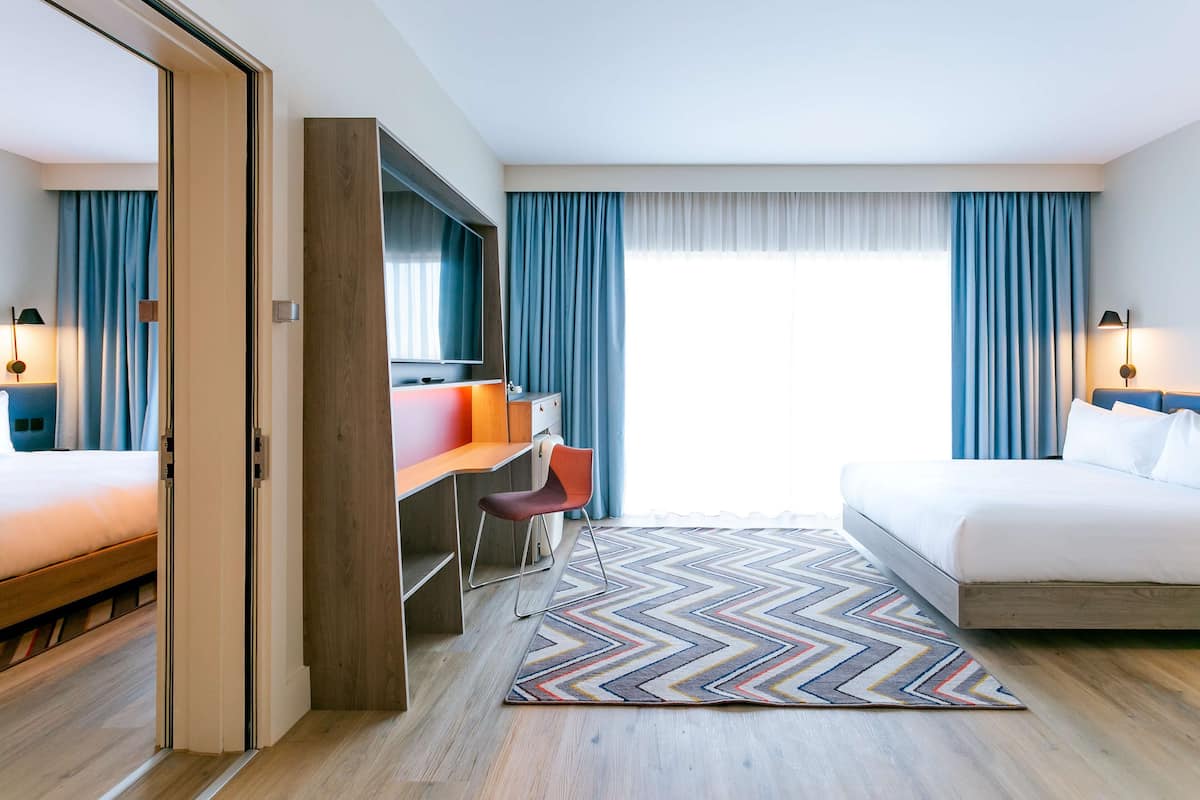 A hotel room with a bed, a desk with a chair, a television, and a large window with curtains.