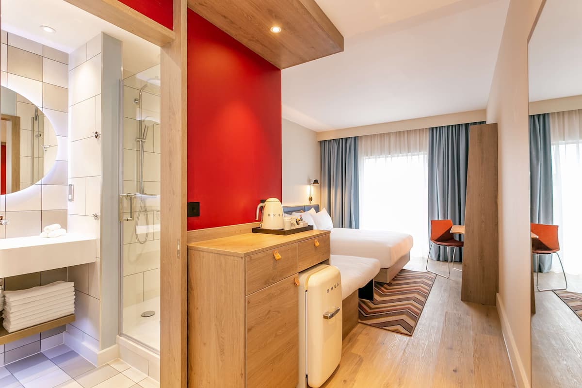 A hotel room with a shower, a bed, a desk with a chair, and a bathroom with a mirror and sink.