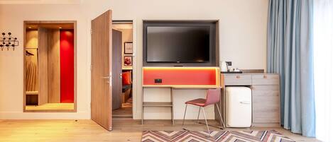 Una habitación de hotel moderna con un televisor de pantalla plana grande, una kitchenette compacta, un vestidor con iluminación roja y una alfombra con estampado en zigzag.