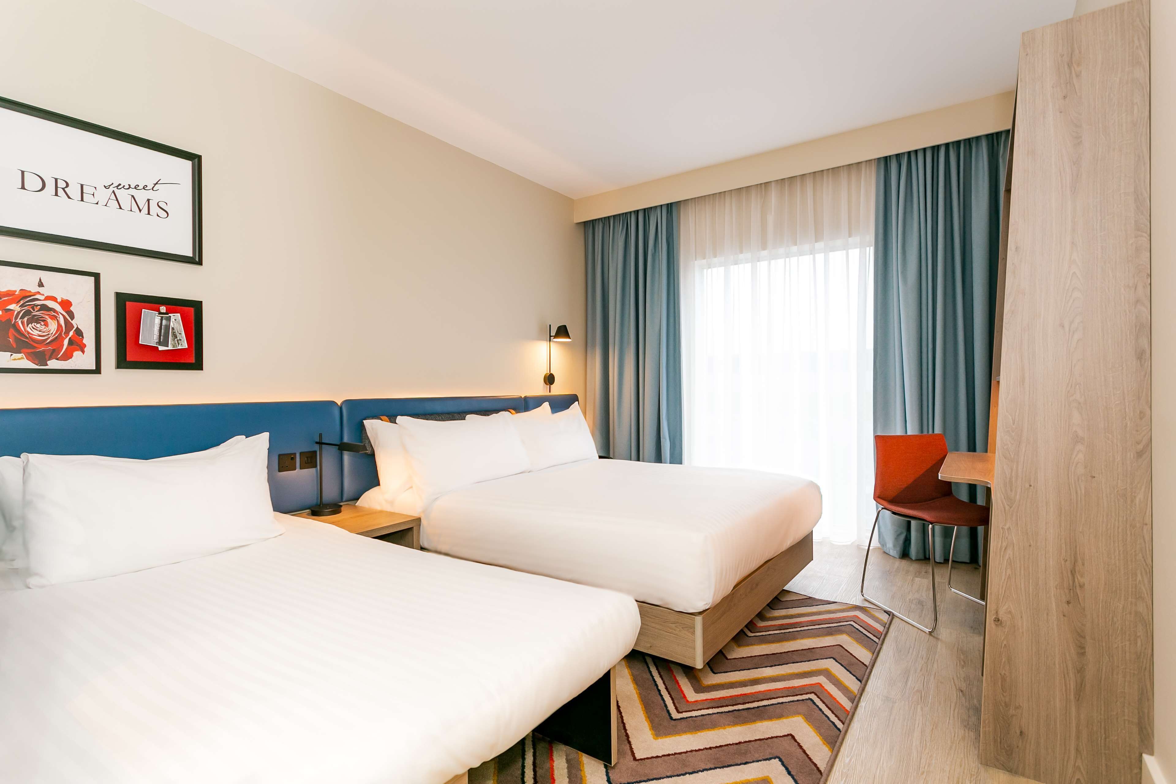 A hotel room with two beds, a desk, a chair, a window with curtains, and framed pictures on the wall.