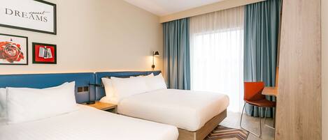 A hotel room with two beds, a desk, a chair, a window with curtains, and framed pictures on the wall.
