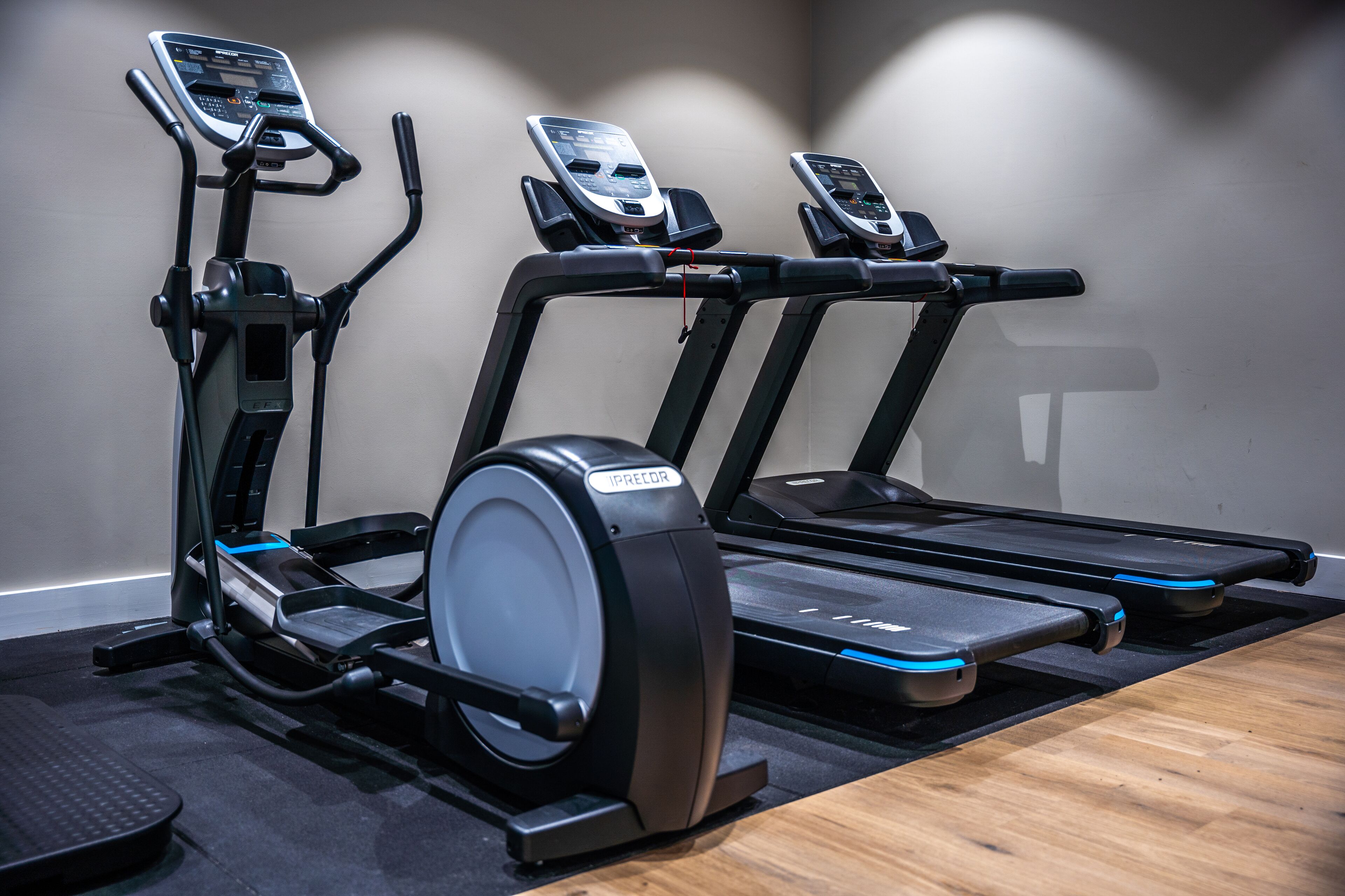 Three stationary bikes and a treadmill in a gym with a wooden floor and a plain wall.