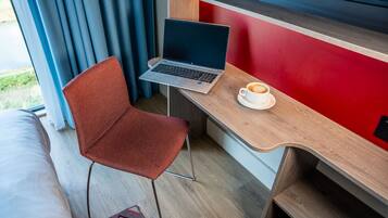 A modern room with a laptop on a wooden desk, a red accent wall, and a view of greenery outside.