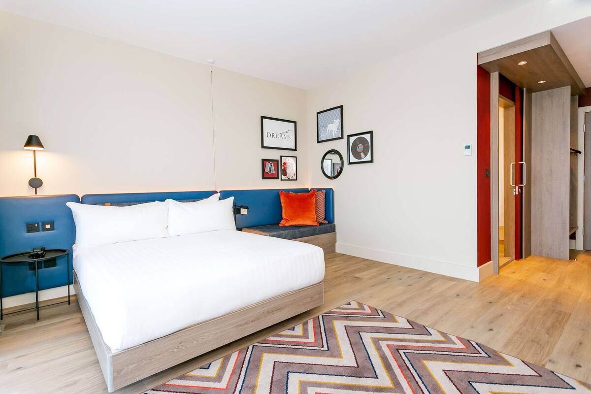 A modern hotel room with a large bed, a bedside table, a lamp, and a wall with framed pictures.