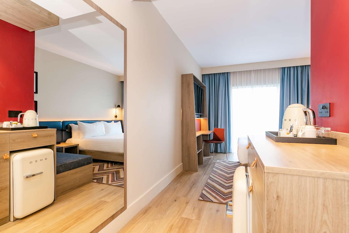 A hotel room with a bed, a desk, a chair, a small fridge, and a window with curtains.