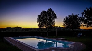 Une piscine rectangulaire bordée de briques, entourée de pelouse et d’arbres, avec un coucher de soleil en arrière-plan.