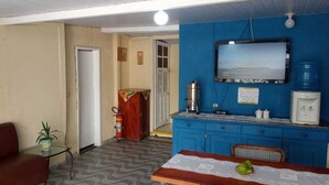 A room with a brown sofa, a small table with a placemat, a blue cabinet with a television, and a water dispenser. - Aldeia Produções Hostel & Bar (Búzios)