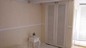 A room with a white tiled floor, a white wall, and a white door with louvered shutters. There is a small white table with a vase and a plant on it. - Aldeia Produções Hostel & Bar (Búzios)